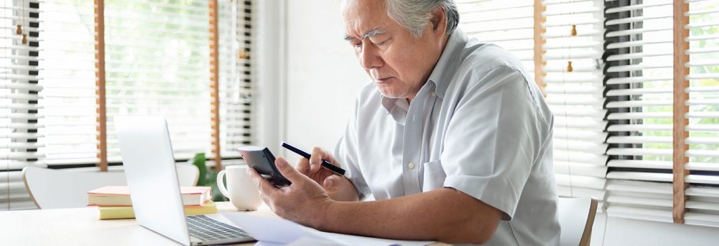 Older man, looking concerned, sitting in his home office, with a laptop and calculator, holding a pen. There's a paperwork on a table.